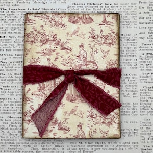 May include: A decorative gift box wrapped in cream-colored paper with a burgundy ribbon. The box is adorned with a vintage-style toile pattern featuring figures and birds, set against a cream background.