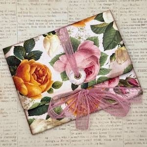 May include: A rectangular paper envelope featuring a floral print with large yellow and pink roses, green leaves, and white buds. The envelope is secured with a pink mesh ribbon tied in a bow and a circular button closure at the center flap.