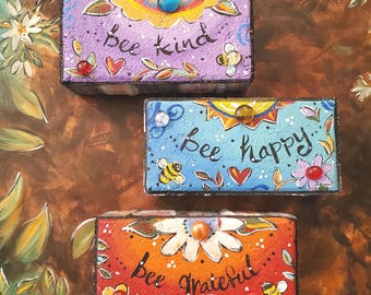 May include: Three wooden boxes with painted floral designs and bee motifs. The boxes are stacked on top of each other. The top box says "bee kind", the middle box says "bee happy", and the bottom box says "bee grateful".