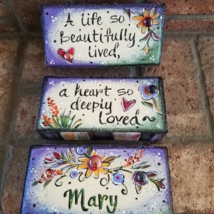 May include: Three painted wooden blocks with a message about life, love, and a name. The top block is green and says "A life so beautifully lived,". The middle block is multi-colored and says "a heart so deeply loved". The bottom block is purple and says "Mary".