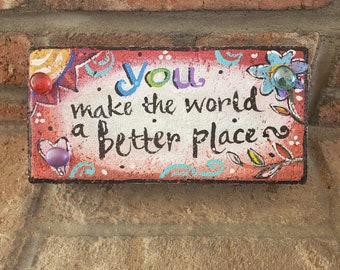May include: A pink and white painted brick with the words "You make the world a better place" in black paint. The brick has colourful painted flowers and a purple heart with a glass gem in the centre.