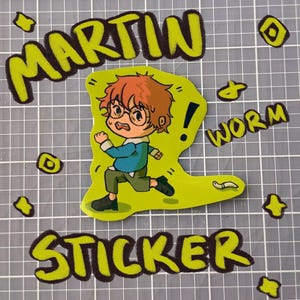 May include: A sticker with a cartoon illustration of a boy with red hair and glasses running. The sticker has the words "MARTIN WORM STICKER" in a green, outlined font. The boy is wearing a blue shirt and green trousers.
