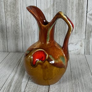 May include: A ceramic pitcher with a glossy finish, featuring a rounded body and a curved handle. The glaze is a blend of deep brown, tan, and vibrant red, with subtle streaks of green throughout the surface. The pitcher has a wide, flared spout.