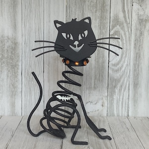 May include: A black metal cat figurine featuring a coiled spring body, a flat head with cut-out eyes and nose, long wire whiskers, and a collar adorned with small orange circular accents. A small white fish skeleton charm hangs from the spring.