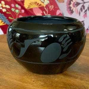 May include: A glossy black ceramic bowl featuring a matte gray bear paw print design encircling the exterior. The bowl has a rounded shape with a wide opening and a smooth, reflective surface finish that contrasts with the muted gray graphic elements.