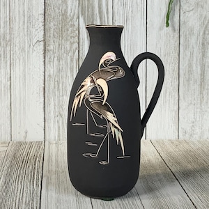 May include: A black ceramic pitcher with a handle and a decorative design of two birds. The birds are depicted in shades of brown and beige, with a touch of pink on their heads. The pitcher has a smooth, matte finish.