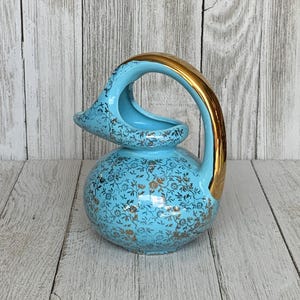 May include: A light blue ceramic pitcher featuring a rounded body and a curved handle with a metallic gold finish. The exterior surface is decorated with a dense, intricate pattern of small gold floral motifs covering the entire blue base.