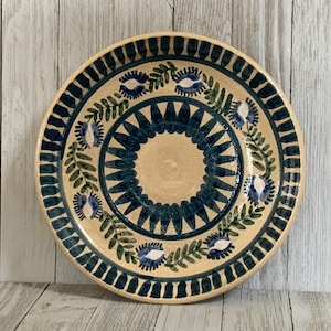 May include: A round ceramic plate with a cream-colored base featuring a dark blue and green hand-painted pattern. The design includes a central sunburst motif, a ring of stylized floral elements, and a dark blue striped border along the edge.