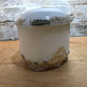 May include: White ceramic jar with a lid, featuring a decorative floral design in brown and green. The jar has a rounded body and a wide, flat lid.