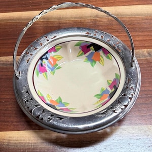 May include: A vintage silver-plated serving basket with a handle. The interior features a cream-colored bowl with a floral design in shades of pink, orange, blue, and green. The rim of the basket has an openwork design.