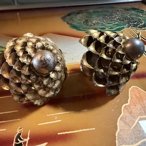 May include: Two decorative objects shaped like pinecones with a metallic gold finish and a smooth, dark, spherical accent at the top of each. The structures feature layered, petal-like scales that create a textured, rounded appearance.