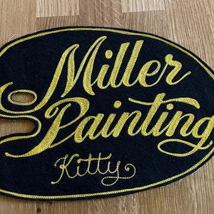 May include: A black felt patch with gold embroidered lettering that reads "Miller Painting Kitty".