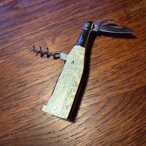 May include: A multi-tool wine opener with a bottle-shaped design. The opener features a corkscrew, knife, and bottle opener. The handle has a pearlescent finish, and the metal components are silver. The tool is set against a wooden surface.