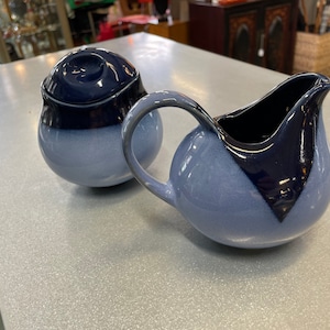 May include: A blue and white ceramic creamer and sugar bowl set. The creamer has a curved handle and a blue rim. The sugar bowl has a lid with a blue knob.
