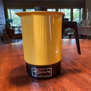 May include: A yellow West Bend Hot Pot Heat & Server with a black handle and base. The hot pot is on a wooden table.