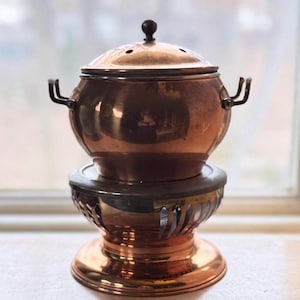 May include: A copper fondue pot with a lid and two handles, sitting on a decorative stand. The pot has a dark knob on top and is displayed on a light-colored surface near a window.