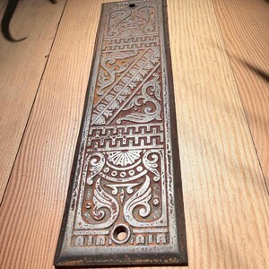 May include: A rectangular metal door push plate featuring a silver-toned finish with raised, intricate scrollwork and geometric patterns. The surface shows signs of oxidation and rust within the recessed areas, and it includes two circular screw holes.