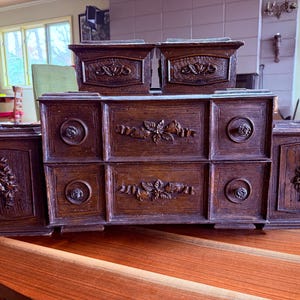 May include: A dark brown wooden dresser with intricate carvings. The dresser has multiple drawers and compartments, with floral and circular designs. The piece is set on a round wooden table.