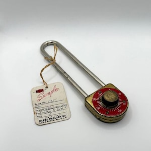 May include: Vintage padlock with a red and gold dial, featuring a combination lock. The lock has a silver-colored shackle and a tag that reads "Sample" and "Reese Padlock Co."