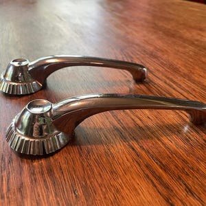 May include: Two chrome-plated door handles. Each handle has a curved design with a fluted base and a rounded top. The handles are set against a wooden surface.