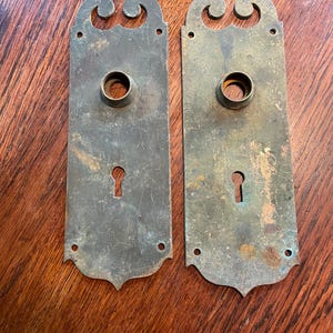 May include: Two antique brass door plates with keyholes and decorative scrollwork at the top. The rectangular plates have a tarnished, aged appearance, showing signs of wear and use. The plates are likely for a vintage or historical door.