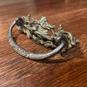 May include: Antique brass drawer pull with a decorative ornate design featuring a stylized face and floral accents.