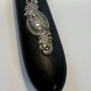 May include: Close-up of a black wooden handle with a decorative silver-colored metal embellishment. The metal piece has an ornate, swirling design. The handle is smooth and rounded.