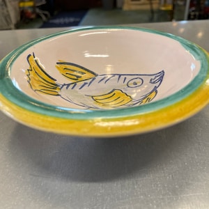 May include: A white ceramic bowl with a green and yellow rim. The bowl features a hand-painted design of a blue and yellow fish.