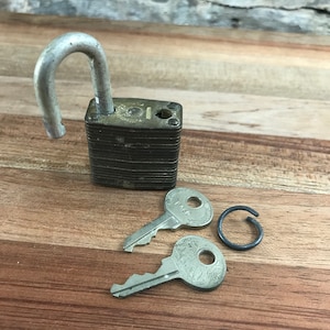 May include: A vintage padlock with a silver-toned body and a silver-toned shackle. Two keys and a metal ring are also included.
