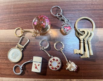 May include: A collection of keychains on a wooden surface. The keychains include a clear orb with pink stars, a small square with a red design, a coin, a lighter with a football player, a round pendant with red gems, and a set of brass keys.