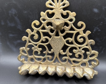 May include: Ornate brass wall sconce with a decorative, openwork design. The sconce features a central shield motif and a scalloped base. The brass has a warm, aged patina, suggesting vintage or antique origins. The overall shape is triangular, with a floral-inspired top.