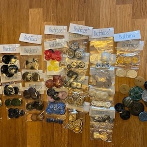 May include: Assortment of buttons in various colors, shapes, and sizes, including black, white, red, blue, green, and yellow. The buttons are packaged in clear plastic bags labeled "Buttons".