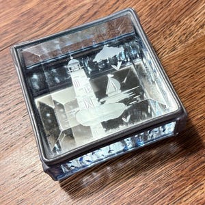 May include: A square glass trinket box with a metal frame and a beveled lid. The lid features an etched design of a lighthouse, a sailboat, and clouds. The sides of the box are made of textured, clear glass panels, providing a decorative container.
