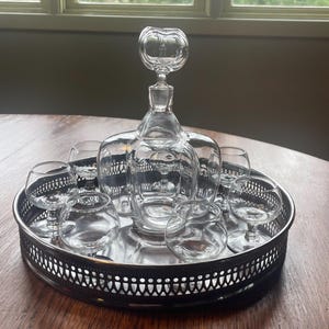 May include: A clear glass decanter with a unique, bulbous stopper, and six matching glasses, all arranged on a silver tray with a decorative border.