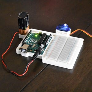 May include: A green circuit board with a USB port and a power jack. A white breadboard is connected to the circuit board. A blue servo motor is connected to the breadboard with orange wires. A battery pack is connected to the circuit board with red and black wires.