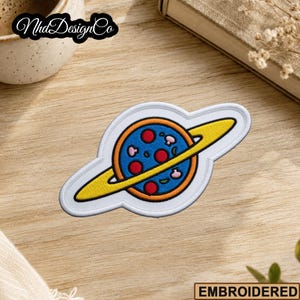 May include: Embroidered patch featuring a pizza planet design. The patch has a blue planet with red pepperoni and mushroom toppings, a yellow ring, and a white border. The text "NhdDesignCo" is in the upper left corner.