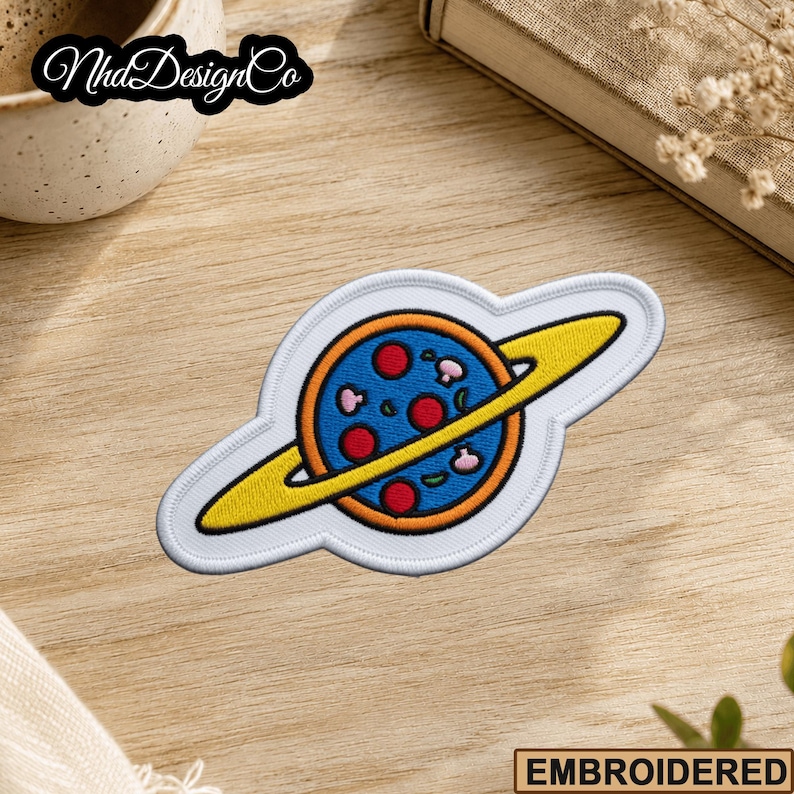 May include: Embroidered patch featuring a pizza planet design. The patch has a blue planet with red pepperoni and mushroom toppings, a yellow ring, and a white border. The text "NhdDesignCo" is in the upper left corner.