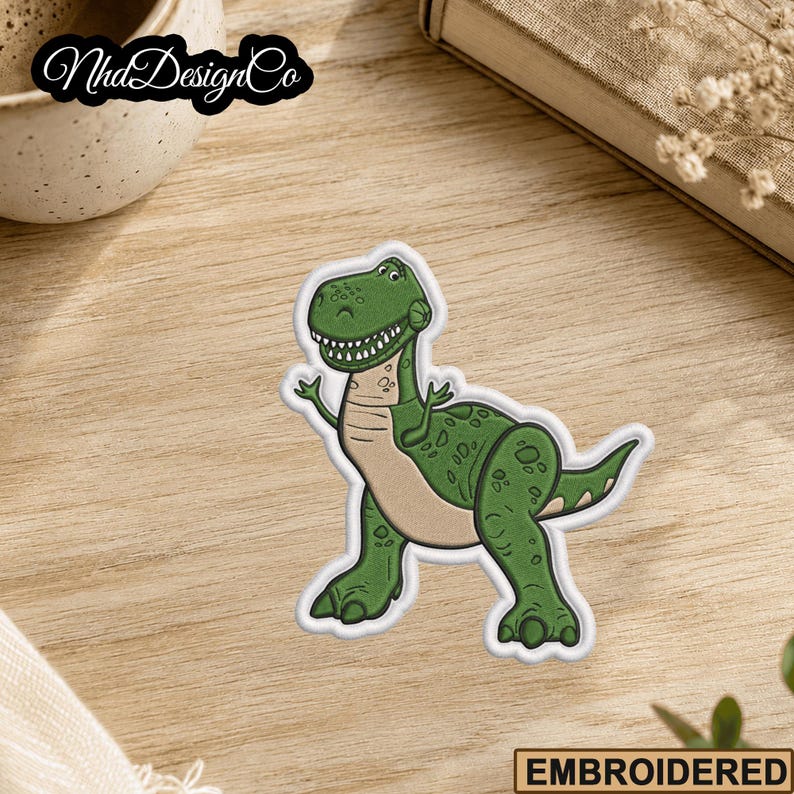 May include: Embroidered patch featuring a cartoon Tyrannosaurus Rex in shades of green and tan. The dinosaur is smiling and waving. The patch has a white border and the text "EMBROIDERED" at the bottom.