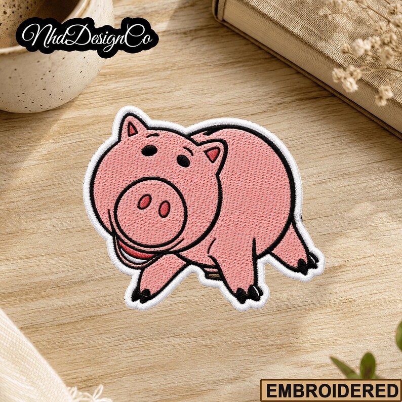 May include: Embroidered patch featuring a pink cartoon pig with black accents. The pig has a smiling expression and is set against a light wooden surface. The patch is outlined in white.