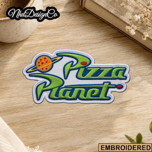 May include: Embroidered patch featuring the words "Pizza Planet" in green and blue, with a planet graphic. The patch has a white border and is on a wooden surface. The word "EMBROIDERED" is at the bottom.