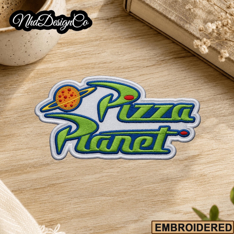 May include: Embroidered patch featuring the words "Pizza Planet" in green and blue, with a planet graphic. The patch has a white border and is on a wooden surface. The word "EMBROIDERED" is at the bottom.