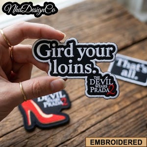 Puede incluir: Parches bordados con el texto "Gird your loins" y "The Devil Wears Prada 2". Los parches son negros con detalles en blanco y rojo. También se ve un parche de zapato de tacón alto rojo.