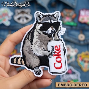Può includere: Toppa ricamata raffigurante un procione con occhiali da sole che beve da una lattina di Diet Coke. Il procione è grigio, nero e bianco, con una coda a strisce. La toppa ha un bordo bianco e la scritta "EMBROIDERED" in un rettangolo marrone.