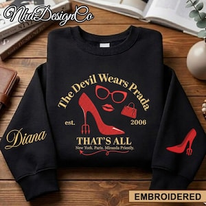 May include: Black sweatshirt with the text "The Devil Wears Prada" and a red high heel, glasses, and purse graphic. The sleeve features the name "Diana" in gold script. The bottom right corner reads "EMBROIDERED".