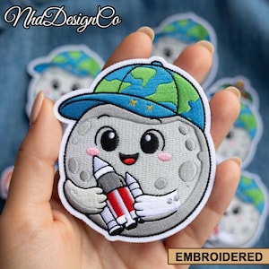 以下が含まれることがあります： 地球をテーマにした青と緑の野球帽をかぶり、ロケットを持っている、笑顔の月のキャラクターが描かれた刺繍パッチ。 パッチは白い縁取りで、「EMBROIDERED」の文字が金の長方形で表示されています。