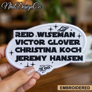 以下が含まれることがあります： 黒い文字でReid、Wiseman、Victor Glover、Christina Koch、Jeremy Hansenの名前が刺繍された白いパッチ。星とロケットのデザインが施された宇宙をテーマにしたパッチです。