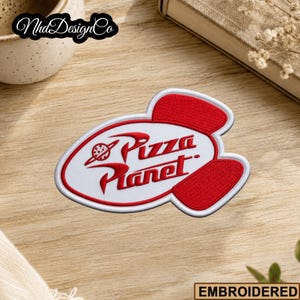 May include: Embroidered patch featuring the Pizza Planet logo in red and white. The patch has a white background with red accents and the words "Pizza Planet" in a stylized font. The patch is on a wooden surface.