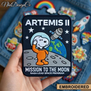 May include: Embroidered patch featuring Snoopy in an orange spacesuit on the moon, with the text "ARTEMIS II" and "MISSION TO THE MOON". The patch includes a satellite, stars, and a depiction of Earth.