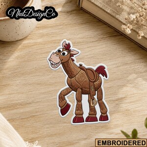 May include: Embroidered patch featuring a cartoon horse with a saddle. The horse is brown with a red mane and tail, and has a smiling expression. The patch has a white border and the word "EMBROIDERED" is at the bottom.