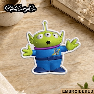 May include: Embroidered patch featuring a green, three-eyed alien character in a blue uniform with a purple collar. The patch has a white border and the text "EMBROIDERED" is at the bottom. The patch is on a wooden surface.